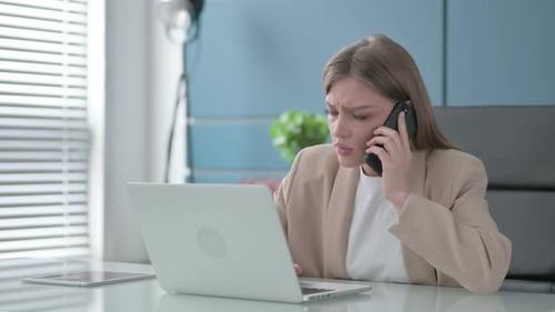 Angry Businesswoman Talking on Smartphone While Using Laptop in Office