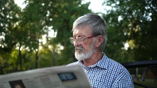 Senior in Plaid Shirt Sits on a Bench in the Park and Reads a Newspaper