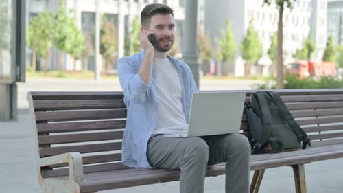 Young Adult Uses Laptop and Phone on Park Bench