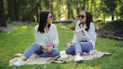 Two Positive Pretty Girlfriends Seemingly Dressed Sitting on a Bedspread on the Grass in the Park