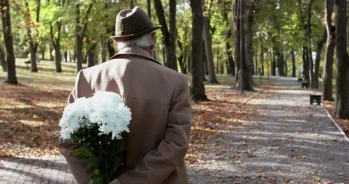 Close Back View of an Elderly Man in a Coat and Hat Walks in Park with Flowers