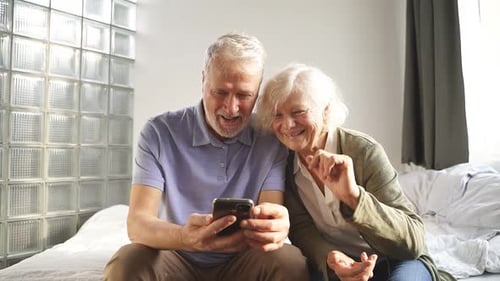 Senior Couple Laughing and Using Smartphone on Bed