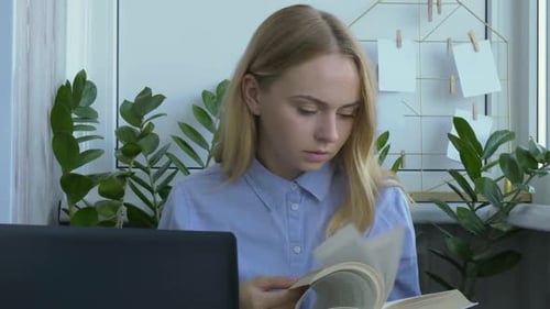 Young Woman Reads Book at Desk with Laptop