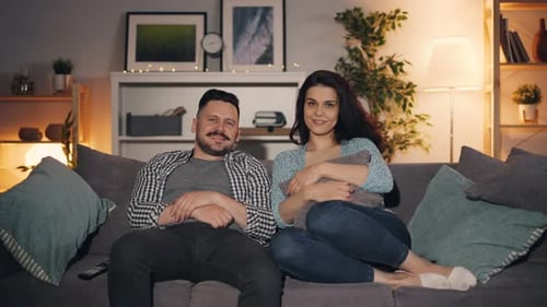 Couple marié regardant la télévision assis sur le canapé dans la maison souriant en regardant la caméra