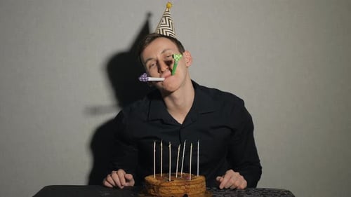 Close Up of Young Man with Cake and a Candles Playing with Party Blowers