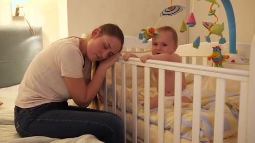 Tired Woman Sleeping While Baby Cries in Crib