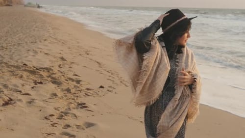 Porträt einer romantischen brünetten Frau mit Hut und Karo, die im Herbst bei Sonnenuntergang am Strand spaziert oder