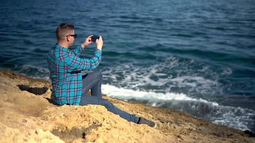 Un joven está sentado con un teléfono en las piedras frente al mar. Hombre con gafas de sol toma fotografías de
