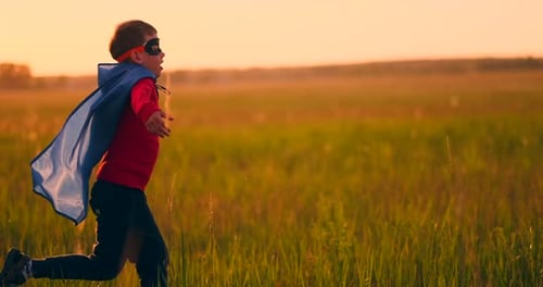 A Boy in a Suit and a Superhero Mask Running Across the Field at Sunset on the Grass