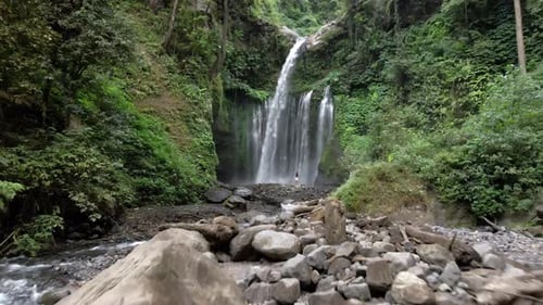 Tiro aéreo para a frente entre colinas cobertas de vegetação e garota na frente da Cachoeira, Lombok.