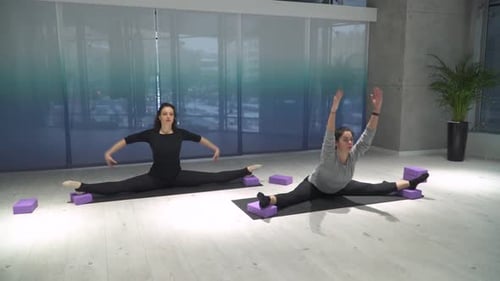 Two Athletic Women Stretching Legs, Leans Forward Sitting on a Splits on Mat in the Gym Near Half