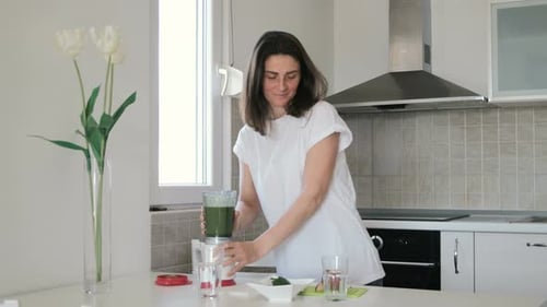 Young woman in white t-shirt prepares green smoothie in blender at home in the kitchen