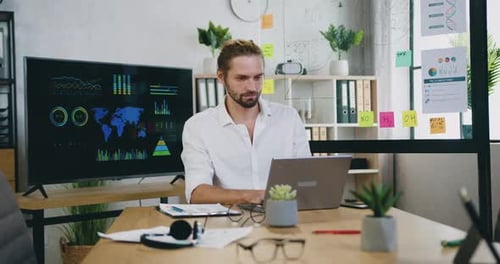 Jeune homme barbu expérimenté et concentré travaillant sur ordinateur dans une salle de travail moderne