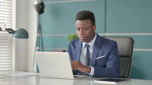 African Businessman Smiling at Camera While Using Laptop in Office