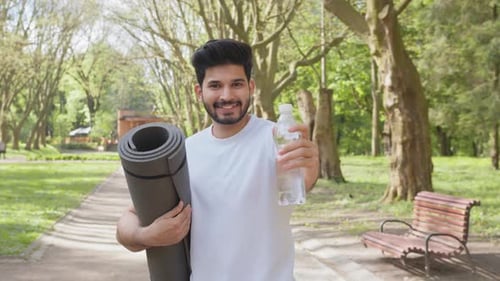 Smiling Man Holding Yoga Mat and Water in Park