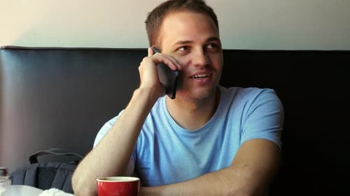 Indoor Shot of Smiling Young Man Sitting and Talking on Mobile Phone. Happy Young Guy at Cafe