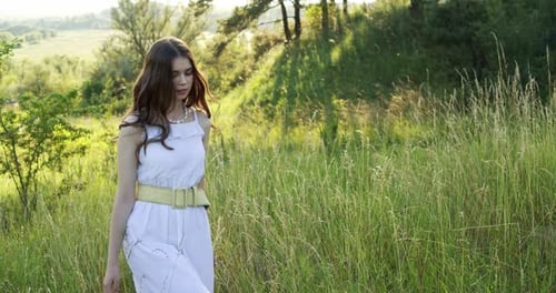 A Cheerful Teenage Girl in Light Dress Walks in a Clearing Between Green Trees in the Summer