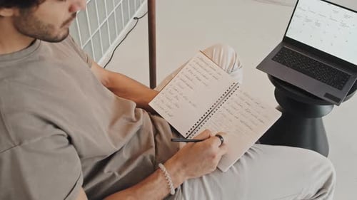 Man Writes in Notebook Next to Laptop Calendar