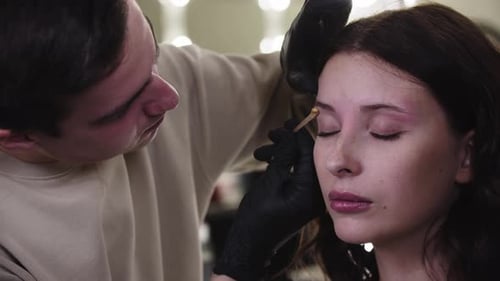 Un maquillador masculino que aplica cera en la parte inferior de la ceja a su bonita clienta en el