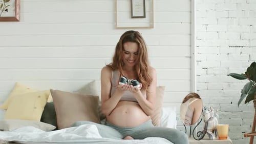 Pregnant Woman Admiring Baby Shoes on Bed