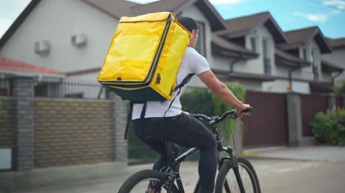 Photo de suivi d'un jeune homme caucasien avec un sac de livraison jaune à vélo dans une rue de banlieue