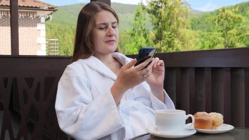 Beautiful Smiling Woman Having Breakfast and Using Smartphone on Hotel Room Balcony
