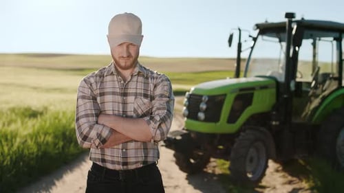 A Young Bearded Farmer is Standing in a Field Near a Tractor