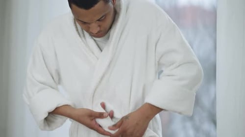 Young Man Tying Bathrobe Belt and Putting Hands in Pockets Standing in Bedroom Indoors