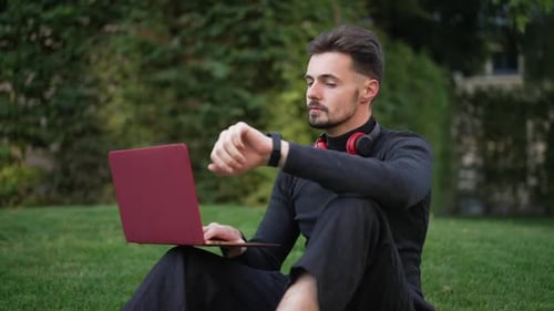 Young Male Freelancer Sitting on Park Meadow Surfing Internet on Laptop Checking Time on Watch