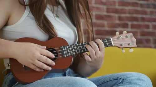 Female Musician is Sitting on Sofa and Playing Ukulele Strumming Strings Front View