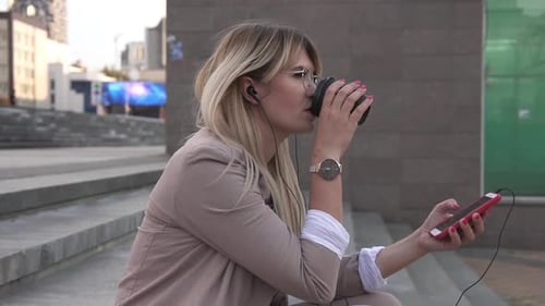 A woman is sitting on the stairs in the center of a big city listening to music.