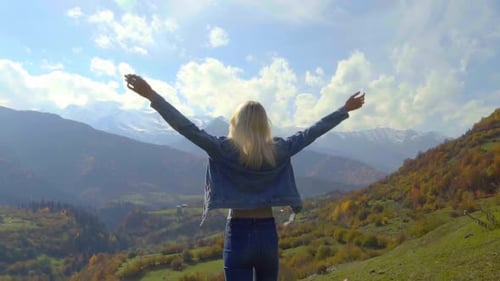woman in denim jacket is standing on top of mountain, raising her hands in air,