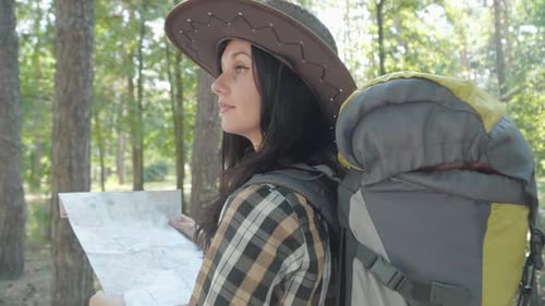 Charming Young Caucasian Female Tourist with Map Looking Around in Sunny Forest and Walking Away