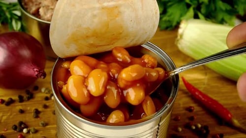 El chef toma con cuchara frijoles guisados en salsa de tomate de una lata de metal sobre una tabla de cocina de madera