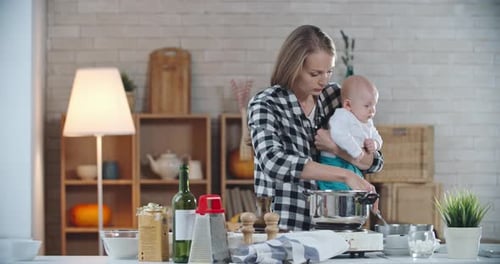 Woman Cooking with Baby in Arm in Kitchen