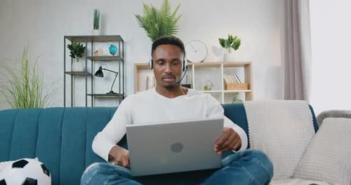 Un mec à la peau noire avec des écouteurs assis sur le canapé à la maison et commençant à travailler sur un ordinateur portable
