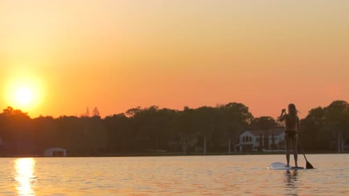 A young woman sup stand-up paddleboarding on a lake at sunset.