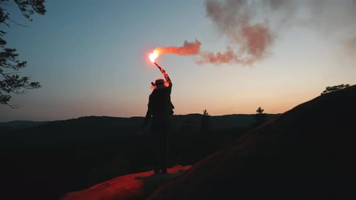 A Young Woman is Standing on the Edge of a Cliff and Waving Flares in the Night