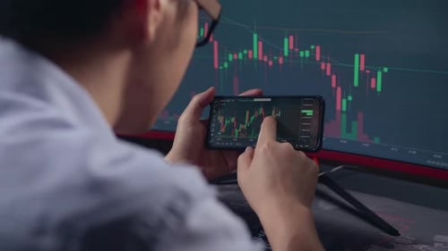 Over The Shoulder View Of Stock Market Trader Working On A Mobile Phone And Computer