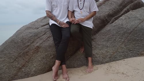 Casal homossexual de mãos dadas sentado perto um do outro em uma grande rocha na praia, sem empurrar o rosto