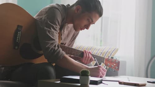 Young Man Plays Guitar and Writes Music Indoors
