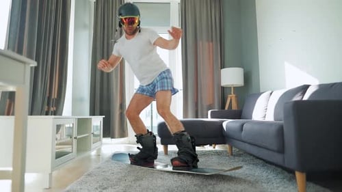 Vídeo divertido. Homem de bermuda e camiseta retrata snowboard em um tapete em um quarto aconchegante. Esperando por