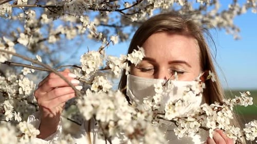 Beautiful girl in a medical mask stands near a flowering tree.