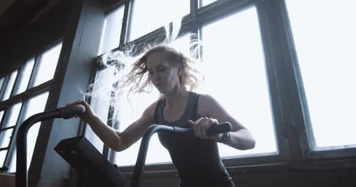 Young Beautiful Athletic Blonde Woman Working Out on Stationary Gym Bike Machine with Wind in Hair