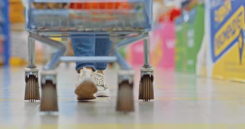 De perto, as pernas de uma mulher de tênis estão andando pelo supermercado carregando um carrinho de compras