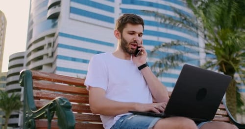 Young Man Sitting in the Park with Palm Trees and Laptop Talking on the Phone