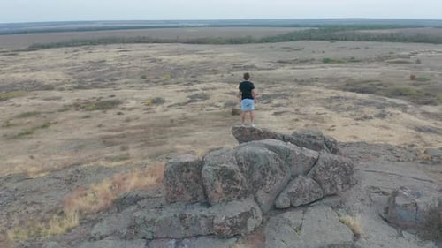 Aerial View. Flying Around Young Man Standing Victoriously on Top of the Mountain at Sunset