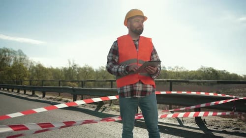 Young Bearded Man Inspector in an Orange Vest and Protective Helmet with Tablet