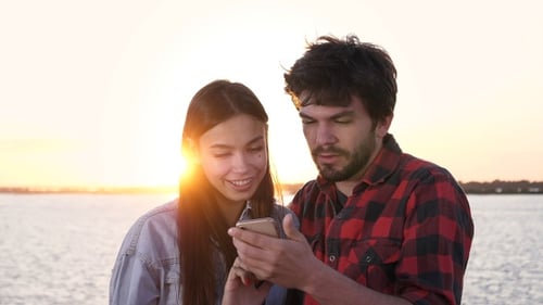 Young Couple Laughing at Phone by Water at Sunset