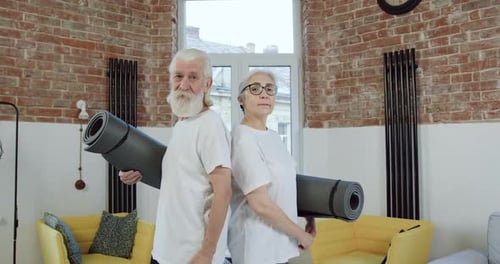 Vieux couple en tenue de sport posant devant la caméra avec des tapis de yoga tout en commençant à faire des exercices à la maison
